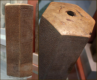 A hexagonal clay prism covered in ancient Akkadian cuneiform script, known as the Taylor Prism, sitting on a museum display base.